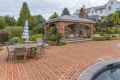 Brick patio featuring a wooden dining set with a blue and white striped umbrella. A covered outdoor fireplace seating area is in the background, enveloped by lush greenery and a stone wall. Experience exquisite outdoor living in Delaware County, PA, with the pool partially visible in the foreground.