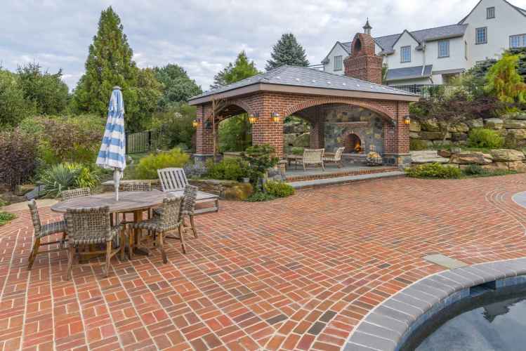 Brick patio featuring a wooden dining set with a blue and white striped umbrella. A covered outdoor fireplace seating area is in the background, enveloped by lush greenery and a stone wall. Experience exquisite outdoor living in Delaware County, PA, with the pool partially visible in the foreground.