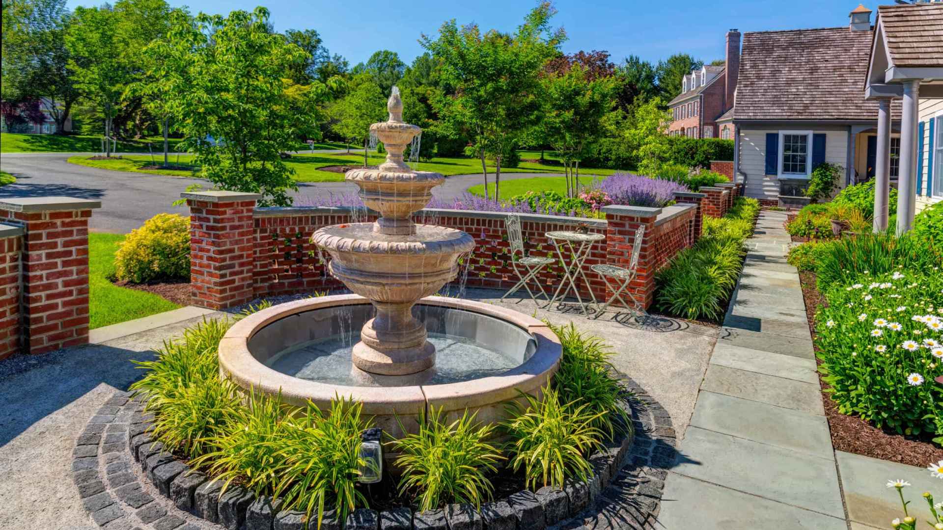 A beautifully landscaped garden, courtesy of premier landscaping services in Delaware County PA, features a stone fountain surrounded by lush greenery. A brick wall encloses the area, where a small patio set invites relaxation. To the right stands a house with a shingled roof under a clear blue sky.