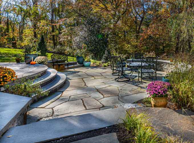 Stone patio with wrought iron furniture, surrounded by colorful potted flowers and greenery. Curved steps lead to a grassy area. Trees with autumn foliage are in the background. Bright sunlight casts shadows across this serene outdoor living space in Delaware County, PA.