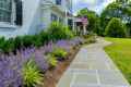 A beautifully landscaped garden beside a white brick house with black shutters showcases outdoor living in Delaware County, PA. Lush greenery and blooming purple flowers line a gray stone pathway. An American flag is displayed near the entrance, with trees standing gracefully in the background.