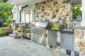 Outdoor kitchen with a stone wall, featuring a stainless steel gas grill, cabinets, and countertop. Nestled within outdoor living in Delaware County, PA, the space is embraced by lush garden blooms and trees, crafting a serene oasis.
