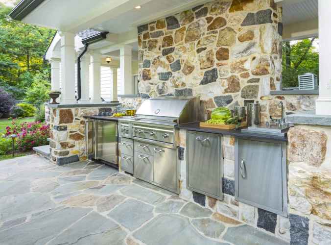 Outdoor kitchen with a stone wall, featuring a stainless steel gas grill, cabinets, and countertop. Nestled within outdoor living in Delaware County, PA, the space is embraced by lush garden blooms and trees, crafting a serene oasis.