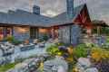 Rustic stone house with a veranda and chimney, warmly lit at dusk. A patio with outdoor seating surrounds a fire pit, perfect for enjoying outdoor living in Delaware County PA. A lush garden and small pond adorned with rocks and greenery decorate the foreground under a cloudy sky.