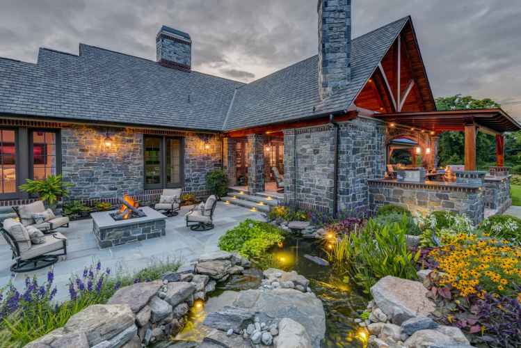 Rustic stone house with a veranda and chimney, warmly lit at dusk. A patio with outdoor seating surrounds a fire pit, perfect for enjoying outdoor living in Delaware County PA. A lush garden and small pond adorned with rocks and greenery decorate the foreground under a cloudy sky.