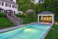 A serene backyard showcases outdoor living in Delaware County, PA, with a lit swimming pool beside a white house. Stone steps lead to a patio with umbrellas and cozy gazebo seating poolside. Lush greenery enhances the tranquil setting, highlighting expert landscaping services in the area.
