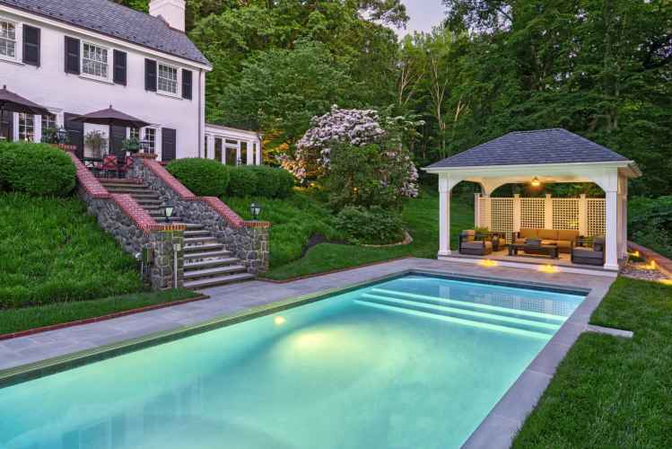 A serene backyard showcases outdoor living in Delaware County, PA, with a lit swimming pool beside a white house. Stone steps lead to a patio with umbrellas and cozy gazebo seating poolside. Lush greenery enhances the tranquil setting, highlighting expert landscaping services in the area.