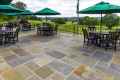 Outdoor patio with stone flooring, a testament to the finesse of hardscaping services in Delaware County, PA. Overlooking lush landscapes, it features tables with dark chairs and large green umbrellas. Trees stretch into a cloudy sky backdrop, embodying serene outdoor living in Delaware County, PA.