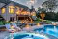 A luxurious house at dusk with glowing windows, overlooking a landscaped backyard crafted by premier landscaping services in Delaware County, PA. Features a wooden deck with stairs, illuminated garden lights, and a stone-lined swimming pool and hot tub. Two lounge chairs sit next to the pool.