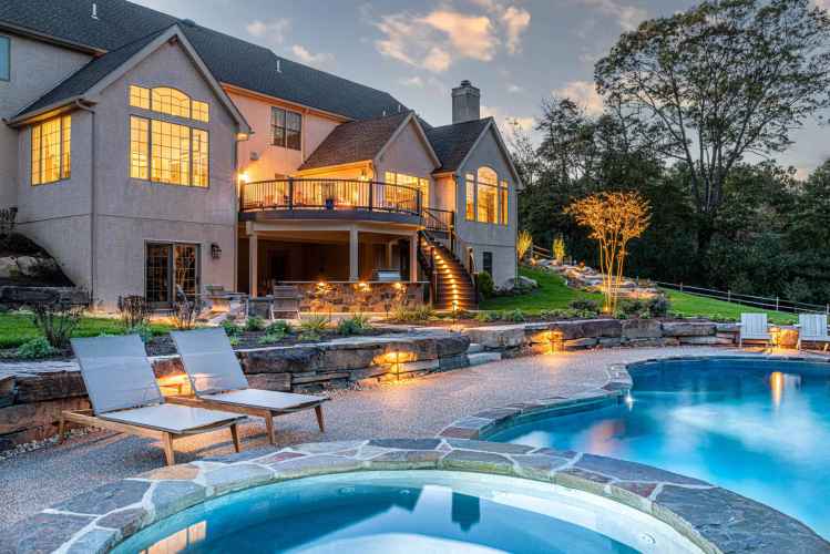 A luxurious house at dusk with glowing windows, overlooking a landscaped backyard crafted by premier landscaping services in Delaware County, PA. Features a wooden deck with stairs, illuminated garden lights, and a stone-lined swimming pool and hot tub. Two lounge chairs sit next to the pool.