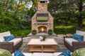 Outdoor living in Delaware County, PA, shines on this patio with wicker couches adorned with blue and white cushions, a wooden coffee table with flowers, and a large stone fireplace. A flat-screen TV shows a sports match as lush greenery envelops the space.