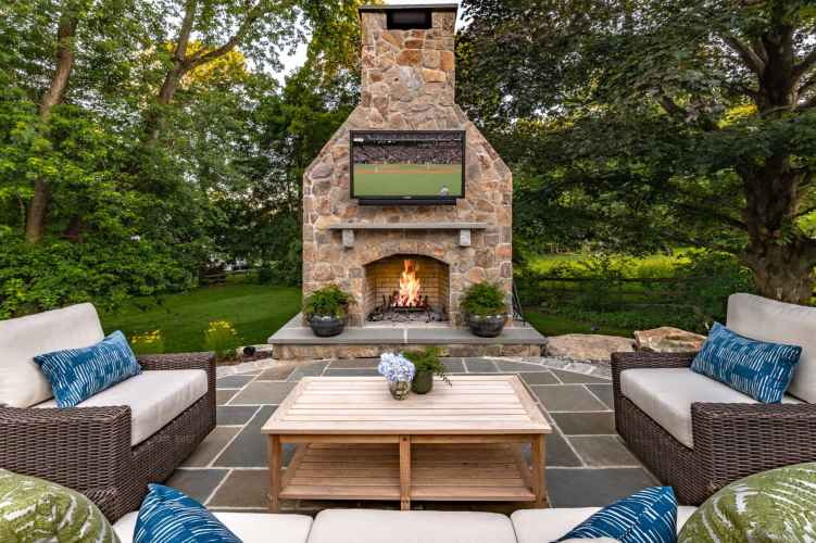 Outdoor living in Delaware County, PA, shines on this patio with wicker couches adorned with blue and white cushions, a wooden coffee table with flowers, and a large stone fireplace. A flat-screen TV shows a sports match as lush greenery envelops the space.