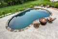 A backyard in Delaware County, PA features an oval swimming pool bordered by stones and surrounded by a paved patio. Two cushioned chairs face a circular fire pit, while lush grass and trees enhance the serene atmosphere. Perfectly crafted with expert hardscaping services to elevate outdoor living.