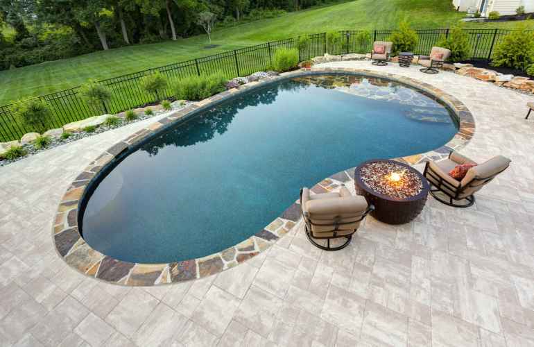 A backyard in Delaware County, PA features an oval swimming pool bordered by stones and surrounded by a paved patio. Two cushioned chairs face a circular fire pit, while lush grass and trees enhance the serene atmosphere. Perfectly crafted with expert hardscaping services to elevate outdoor living.