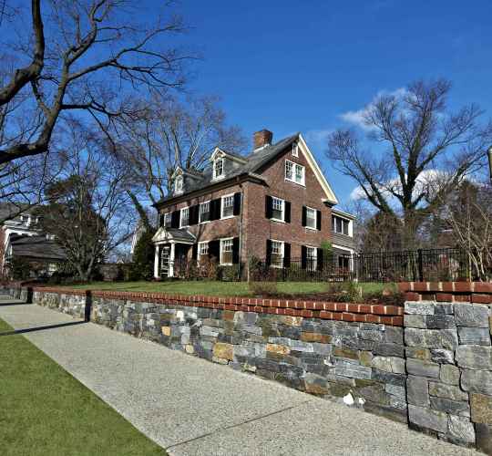 A large brick house with white trim sits behind a low stone wall and a sidewalk, enhanced by professional landscaping services in Delaware County, PA. It features multiple windows, a prominent chimney, and a gabled roof. Leafless trees surround the property under a clear blue sky.