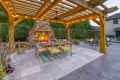 Outdoor living in Delaware County, PA comes alive with a wooden pergola adorned with string lights. A lit stone fireplace and a fire pit add warmth, while the dining table set on a tiled floor with rugs, potted plants, and comfy chairs enhance this beautifully hardscaped scene.