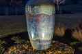 A tall, illuminated ceramic fountain vase sits on a bed of pebbles in a landscaped garden at night, showcasing the artistry of outdoor living in Delaware County, PA. The vase features a textured, multicolored glaze amidst trees and grass under a dark, clear sky.