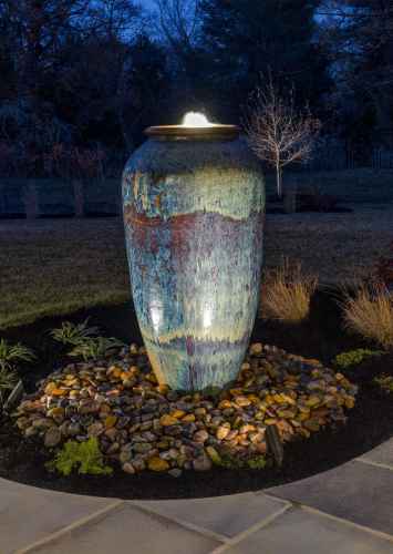 A tall, illuminated ceramic fountain vase sits on a bed of pebbles in a landscaped garden at night, showcasing the artistry of outdoor living in Delaware County, PA. The vase features a textured, multicolored glaze amidst trees and grass under a dark, clear sky.