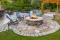 A landscaped backyard in Delaware County, PA, showcases outdoor living at its finest with a stone fire pit and wooden chairs with blue cushions. Behind it, a pool and patio furniture rest beneath a pergola adorned with string lights, all framed by lush greenery.