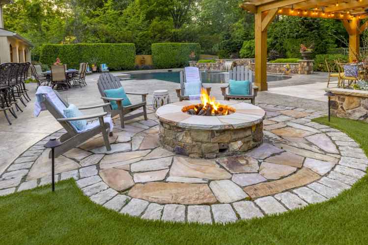 A landscaped backyard in Delaware County, PA, showcases outdoor living at its finest with a stone fire pit and wooden chairs with blue cushions. Behind it, a pool and patio furniture rest beneath a pergola adorned with string lights, all framed by lush greenery.