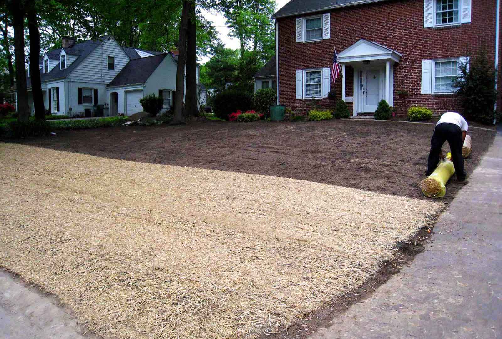 A person unrolls a bale of straw onto a newly cleared front yard of a brick house, showcasing expert landscaping services in Delaware County, PA. Nearby, another home is visible with trees lining both properties. An American flag hangs by the front door of the brick house.