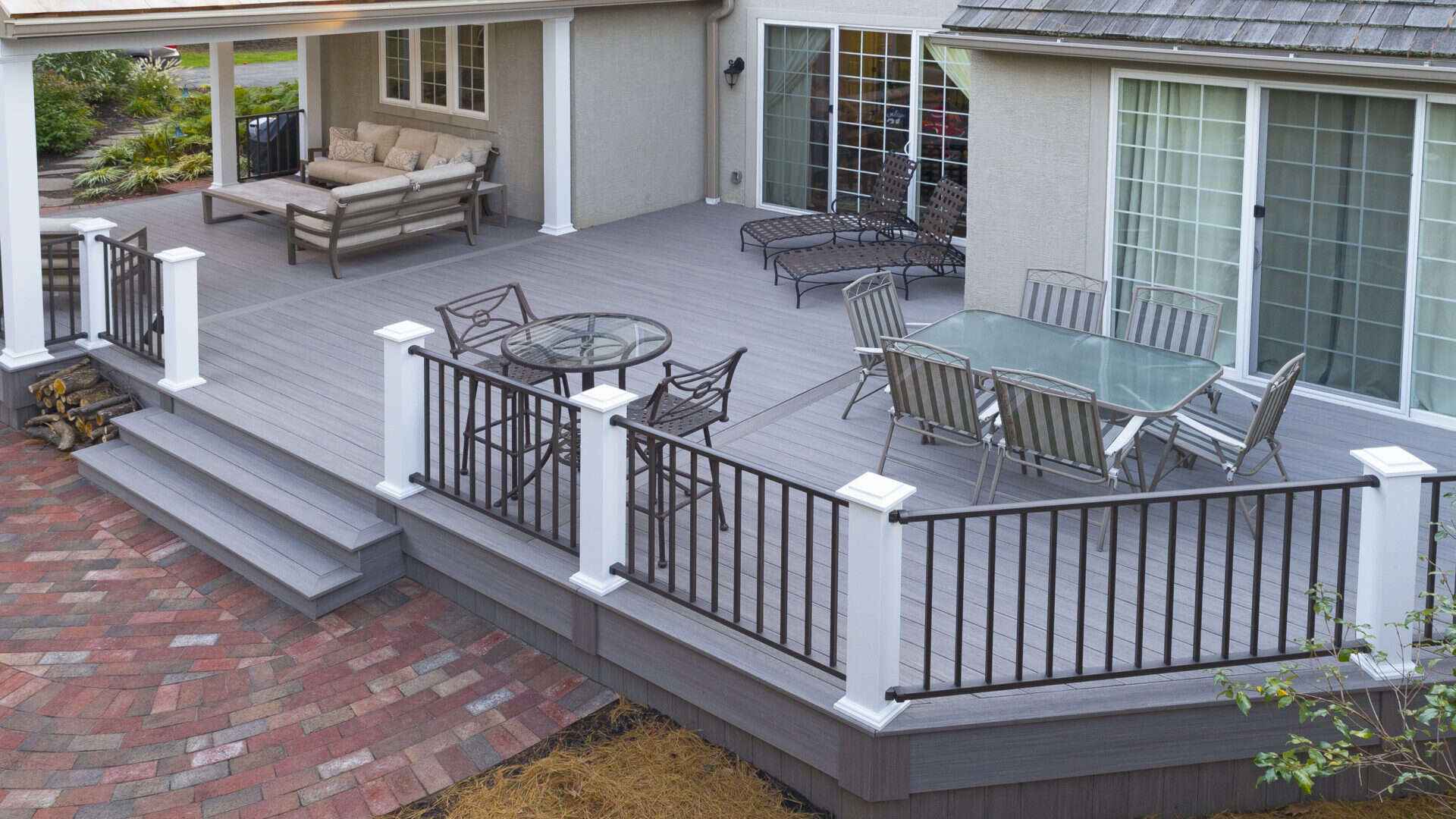 A spacious outdoor deck with gray flooring features two glass-top tables surrounded by metal chairs. A couch and loungers sit near large sliding glass doors. The deck, enhanced by local hardscaping services Delaware County PA, is bordered by white and black railing, overlooking a brick patio below.