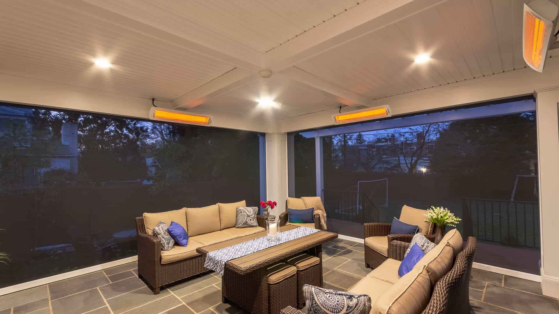 A cozy outdoor patio with wicker furniture, cushioned seats, and blue pillows showcases exquisite outdoor living Delaware County PA style. A table with a blue runner and vase of red flowers is at the center. Overhead heaters are on, and the area is enclosed by screens. Twilight view in the background.