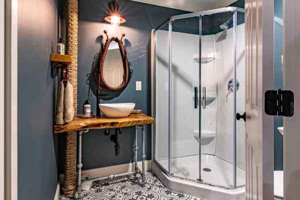 A stylish bathroom with dark blue walls and a patterned tile floor. It boasts a round mirror above a rustic wooden vanity with vessel sink. A glass shower enclosure stands to the right, completing this serene indoor space—a perfect retreat from your outdoor living in Delaware County, PA.
