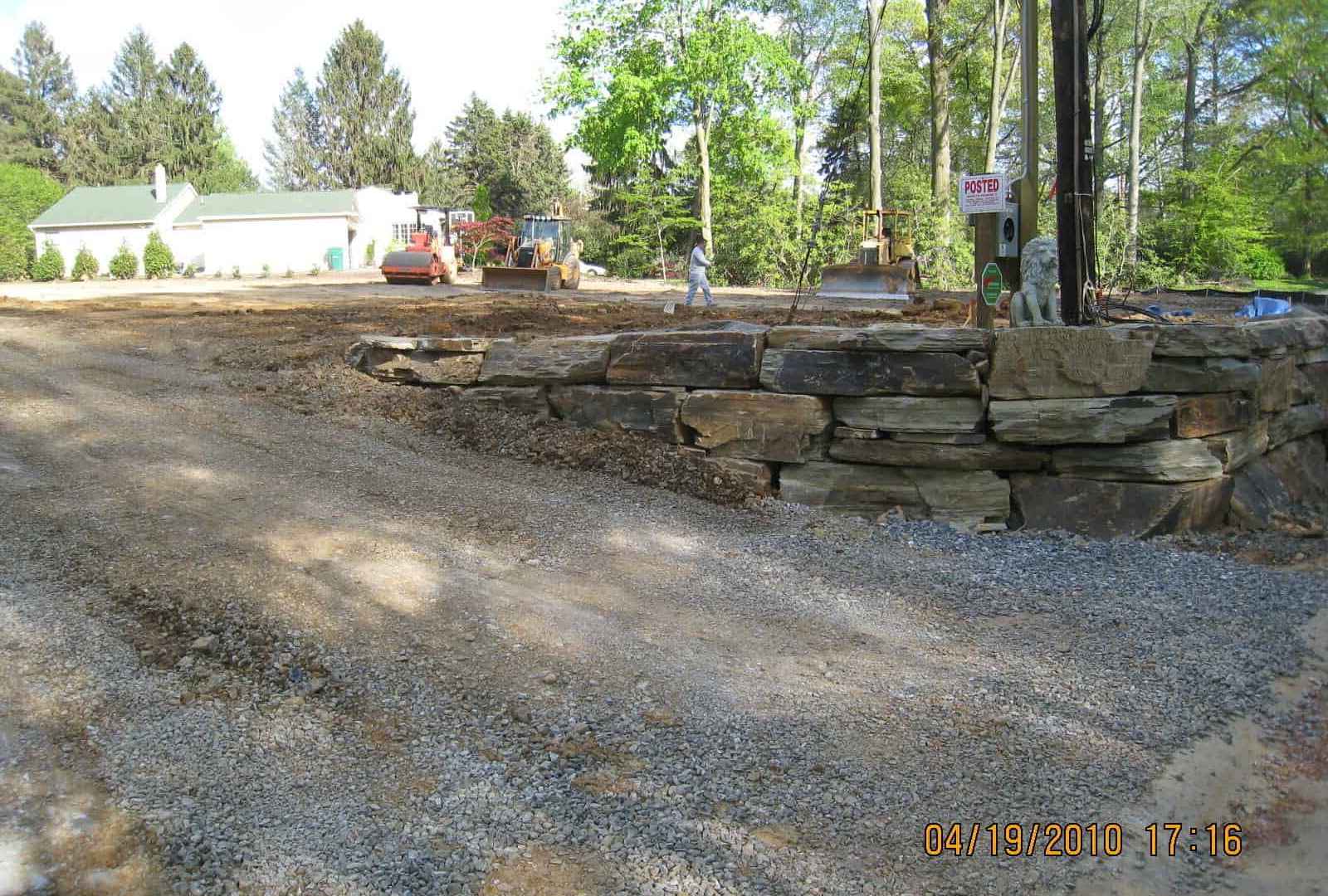 A construction site features a gravel-paved area and a stone retaining wall, perfect for outdoor living in Delaware County, PA. Workers and machinery buzz about, flanked by trees and a white building. A sign reads 