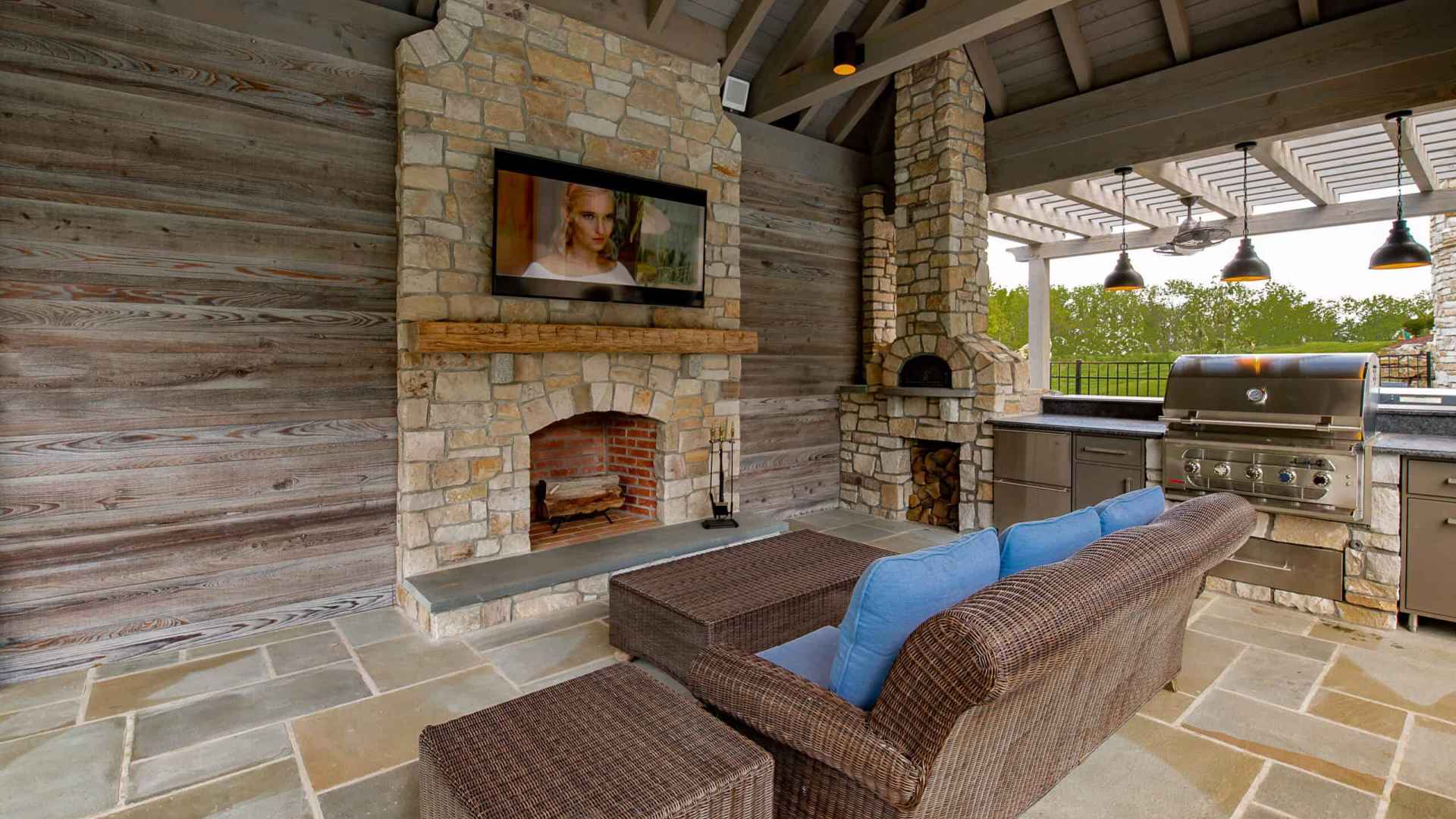 Experience outdoor living in Delaware County, PA, with a stunning patio featuring a stone fireplace and flat-screen TV. Relax on the wicker sofa with blue cushions while enjoying the stainless steel grill station under a pergola adorned with hanging lights. Perfectly designed for comfort and style!.