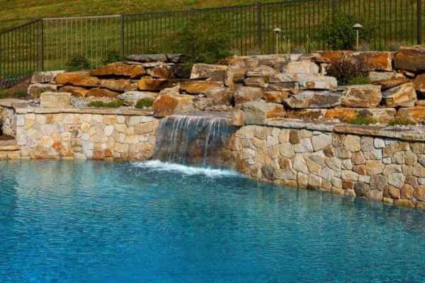 A serene swimming pool with clear blue water and a cascading waterfall feature made of stacked stones, enhanced by expert hardscaping services. The pool is surrounded by a rock wall, with a grassy hillside and metal fence visible in the background, epitomizing outdoor living in Delaware County, PA.