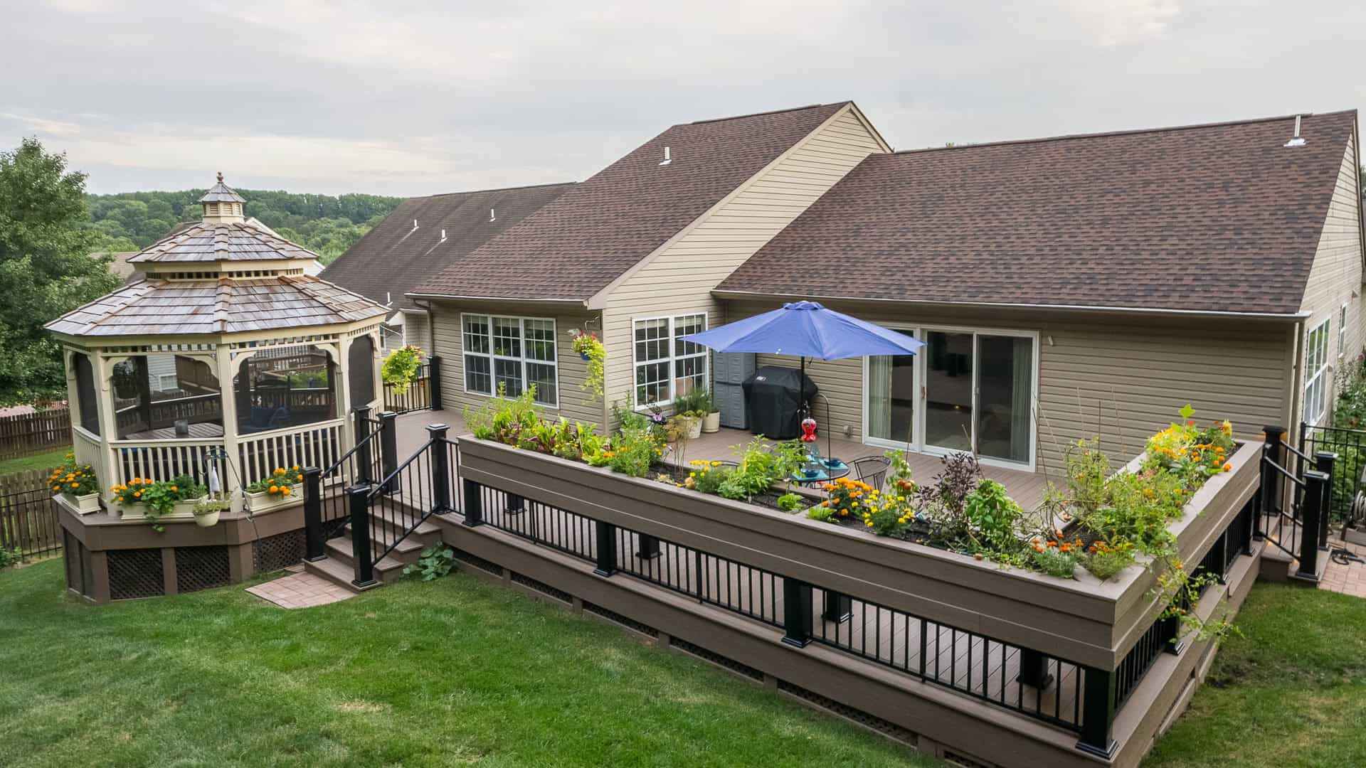 A backyard in Delaware County, PA, showcases a large wooden deck adorned with planters full of vibrant flowers and plants. A blue umbrella offers shade as a child plays nearby, with a charming gazebo to the left. The beige house boasts large windows, highlighting expert landscaping services.