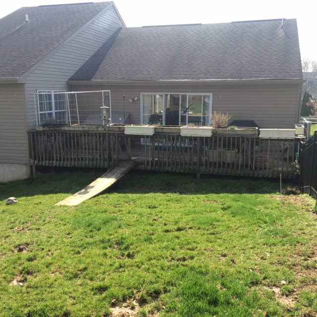A backyard with an elevated deck features a fenced enclosure and planter boxes, perfect for enhancing outdoor living in Delaware County, PA. A ramp extends from the deck to the grass below. The house is beige with a sloped roof, complemented by sliding glass doors leading to the deck.
