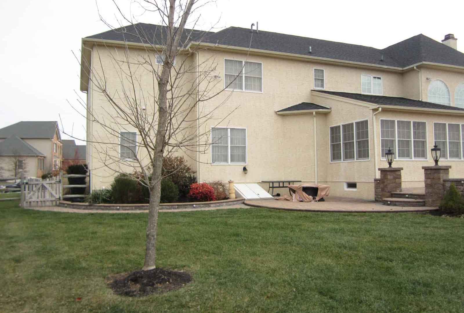 A two-story beige house with a dark roof and manicured lawn invites you into its embrace. A patio with columns offers an ideal space for outdoor living in Delaware County, PA. Bushes and shrubs line the base, while a small tree stands tall against an overcast sky.