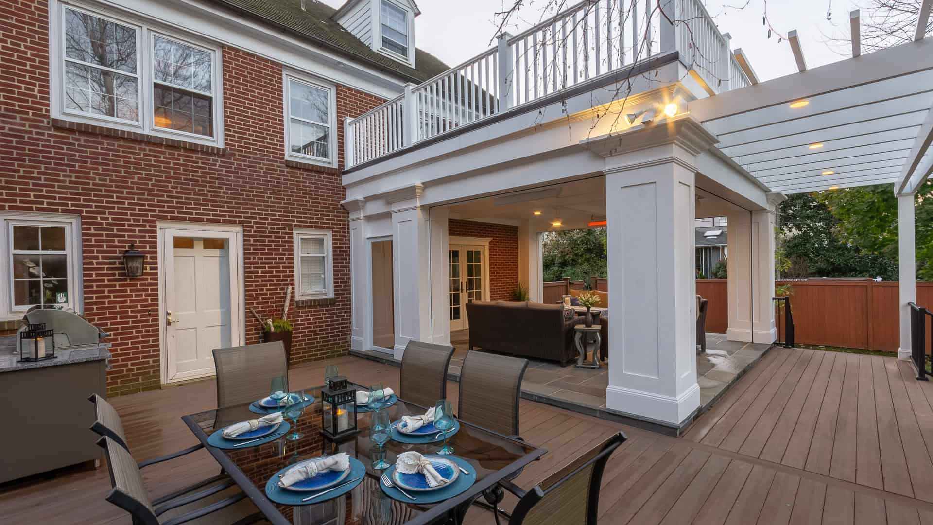 Outdoor living elevated: A wooden deck provides the perfect dining space with a glass table set for dinner, featuring blue napkins and dishware. Attached to a red brick house, it showcases beautiful surroundings. Ideal for those in Delaware County, PA seeking landscaping services.