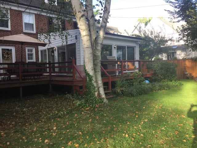 Backyard view of a brick house in Delaware County, PA, featuring a wooden deck with a patio table and umbrella. A large tree stands in the foreground. The grassy lawn, ideal for outdoor living, is dotted with leaves and plants. A fence and neighboring houses complete this charming scene.
