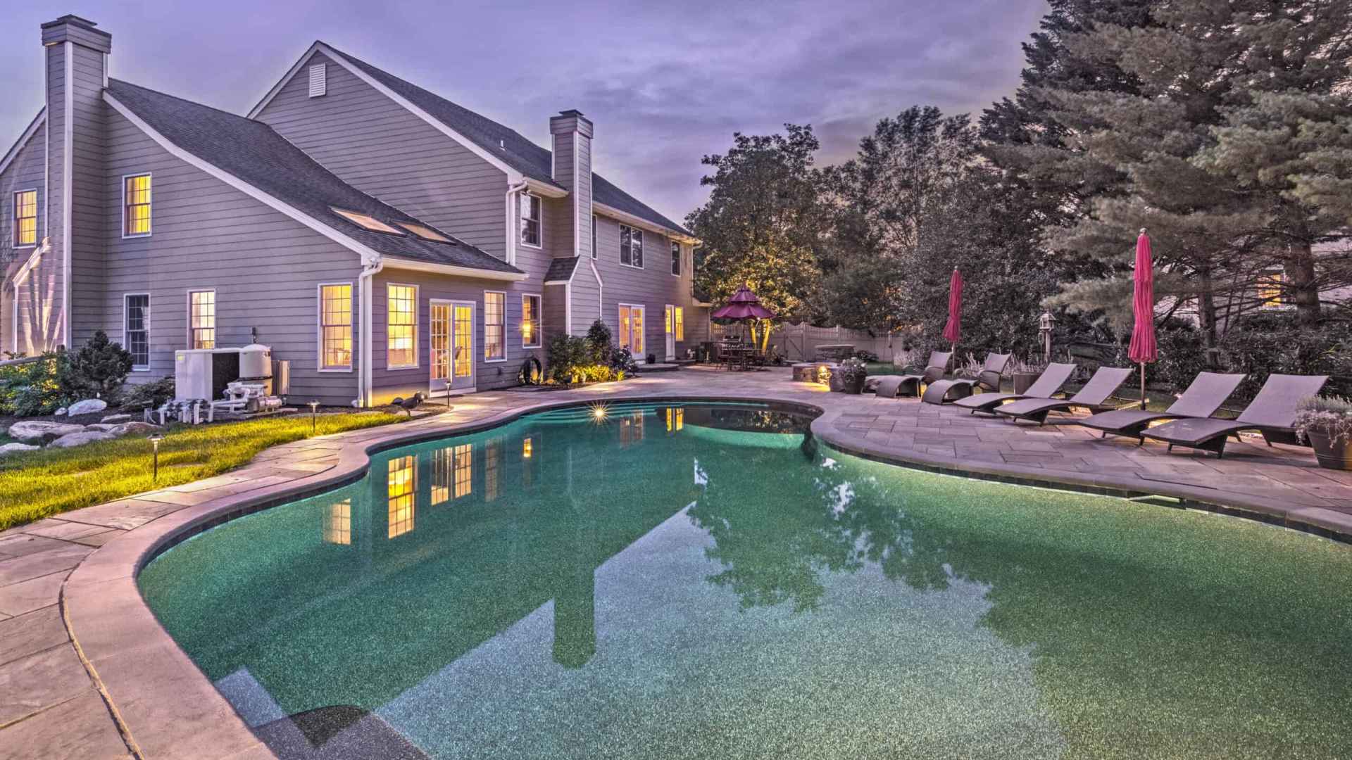 A large backyard in Delaware County, PA, features a pool with gentle lighting. Surrounding the pool are lounge chairs with red umbrellas and lush greenery designed by premier landscaping services. The adjacent two-story house has glowing windows, creating a warm, inviting atmosphere in the evening light.