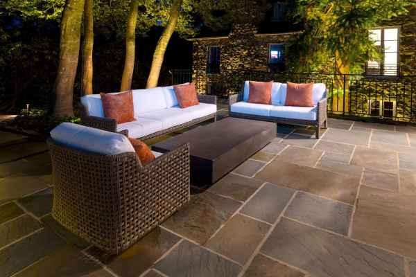 Outdoor living in Delaware County, PA is elevated with this nighttime patio scene. Two wicker sofas and an armchair, adorned with white cushions and brown pillows, encircle a rectangular coffee table on a stone floor. A backdrop of trees and illuminated windows completes the serene setting.