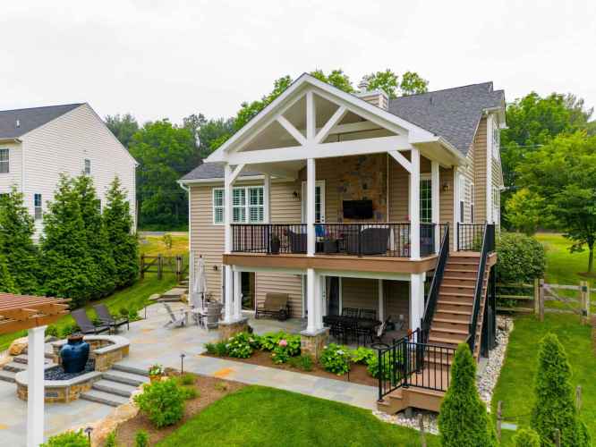 This two-story house boasts a covered patio and deck with outdoor seating, a TV, and decorative plants. Enjoy the stone fire pit surrounded by chairs on the lower patio, all enhanced by expert hardscaping services in Delaware County, PA against a backdrop of lush greenery and trees.