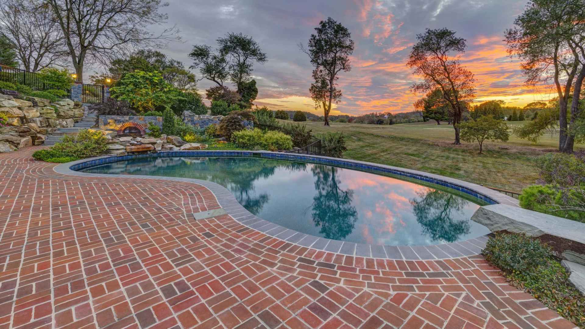 A serene swimming pool bordered by red brick tiles reflects a vibrant sunset sky. Lush greenery and trees, curated through expert landscaping services in Delaware County, PA, surround the pool area, creating a tranquil outdoor setting. The horizon features open fields under a colorful sky with scattered clouds.