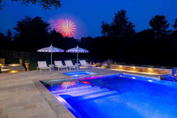 A serene backyard pool in Delaware County, PA, is illuminated with blue lights at night, surrounded by lounge chairs and two umbrellas. Fireworks burst in the sky above, adding a festive touch to this tranquil outdoor living space framed by trees.