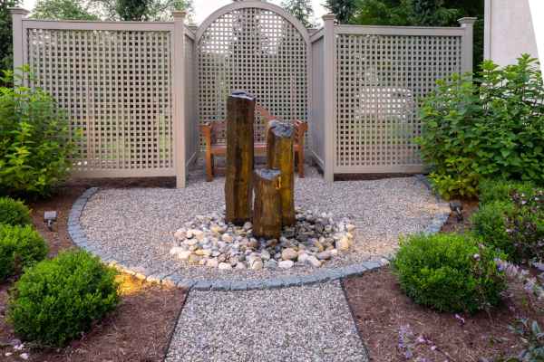 A tranquil garden showcases expert hardscaping services in Delaware County, PA, featuring a gravel path leading to a water feature framed by a wooden trellis and lush plants. Vertical wooden posts form the fountain, while a wooden bench invites relaxation in this serene outdoor living space.