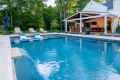 A modern backyard in Delaware County, PA features a swimming pool with a waterfall. A white house with a covered patio, bar seating, and outdoor furniture enhances the outdoor living experience. Expert landscaping services have integrated surrounding trees for a serene, natural setting.