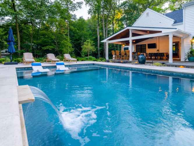 A modern backyard in Delaware County, PA features a swimming pool with a waterfall. A white house with a covered patio, bar seating, and outdoor furniture enhances the outdoor living experience. Expert landscaping services have integrated surrounding trees for a serene, natural setting.