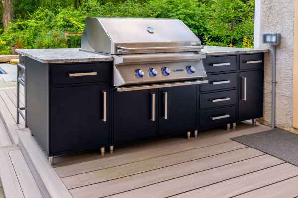 Outdoor living in Delaware County, PA is elevated with this sleek, stainless steel grill on a black cabinet base. Nestled on a wooden deck and surrounded by lush greenery, the grill boasts five control knobs and ample counter space, perfect for any outdoor cooking aficionado.