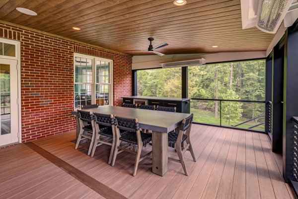 This screened porch offers a charming outdoor living space with wood flooring and ceiling, a large stone dining table encircled by chairs, a ceiling fan, and recessed lighting. Gaze at the serene forest views through expansive windows, all enhanced by expert hardscaping services in Delaware County PA.