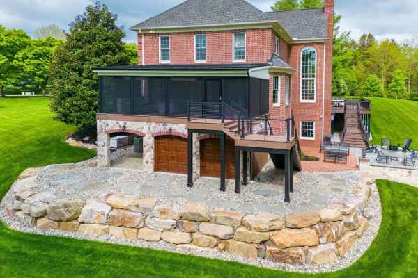A large brick house with a stone foundation, screened porch, and multiple levels features hardscaping services in Delaware County, PA. Enjoy the wooden staircase, spacious patio, and stone-bordered lawn amidst lush grass and surrounding trees under a cloudy sky.