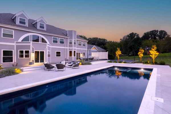 Modern two-story house with gray siding, large windows, and a spacious patio featuring outdoor furniture. A rectangular swimming pool reflects the evening sky, enhanced by expert hardscaping services from Delaware County PA, surrounded by greenery and a well-lit garden area.