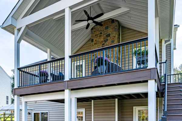 Elevated two-story porch in Delaware County, PA, with a gabled roof and white trusses. Upper level boasts outdoor seating and ceiling fan, while the lower level showcases a patio dining set. Stone and clapboard finish blend seamlessly with expert landscaping services and stairs leading up.