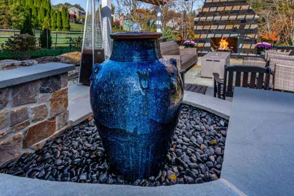 A large blue ceramic vase stands amid smooth black pebbles on a patio in Delaware County, PA. Nearby, an inviting outdoor seating area and a stone fireplace with a lively fire are beautifully integrated with lush greenery, showcasing the charm of expert hardscaping services.